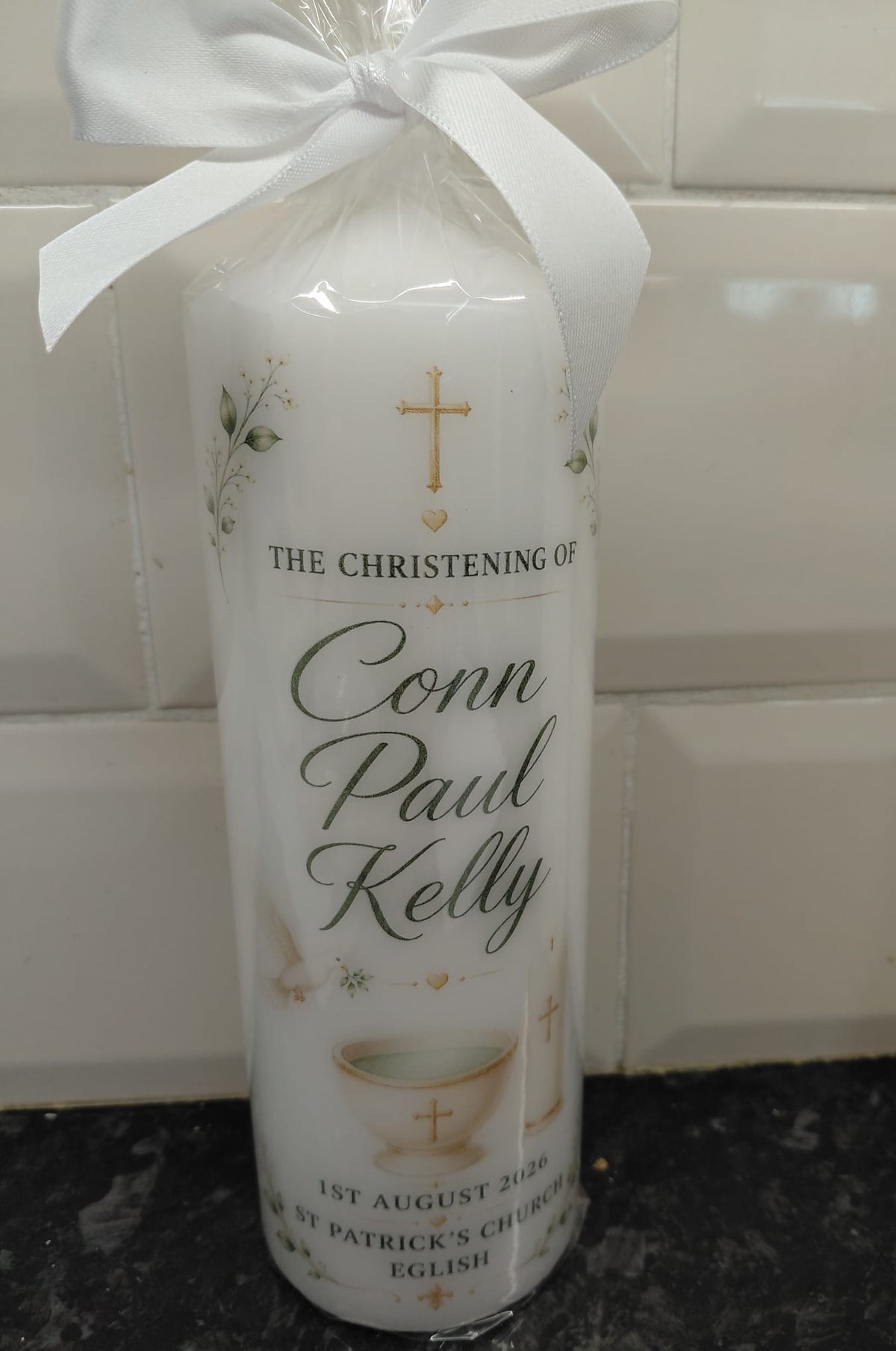 A personalized white pillar candle for a christening, wrapped in clear cellophane with a white satin bow. The label reads 'THE CHRISTENING OF Conn Paul Kelly' with a gold cross and communion cup illustration, set against a tiled background.