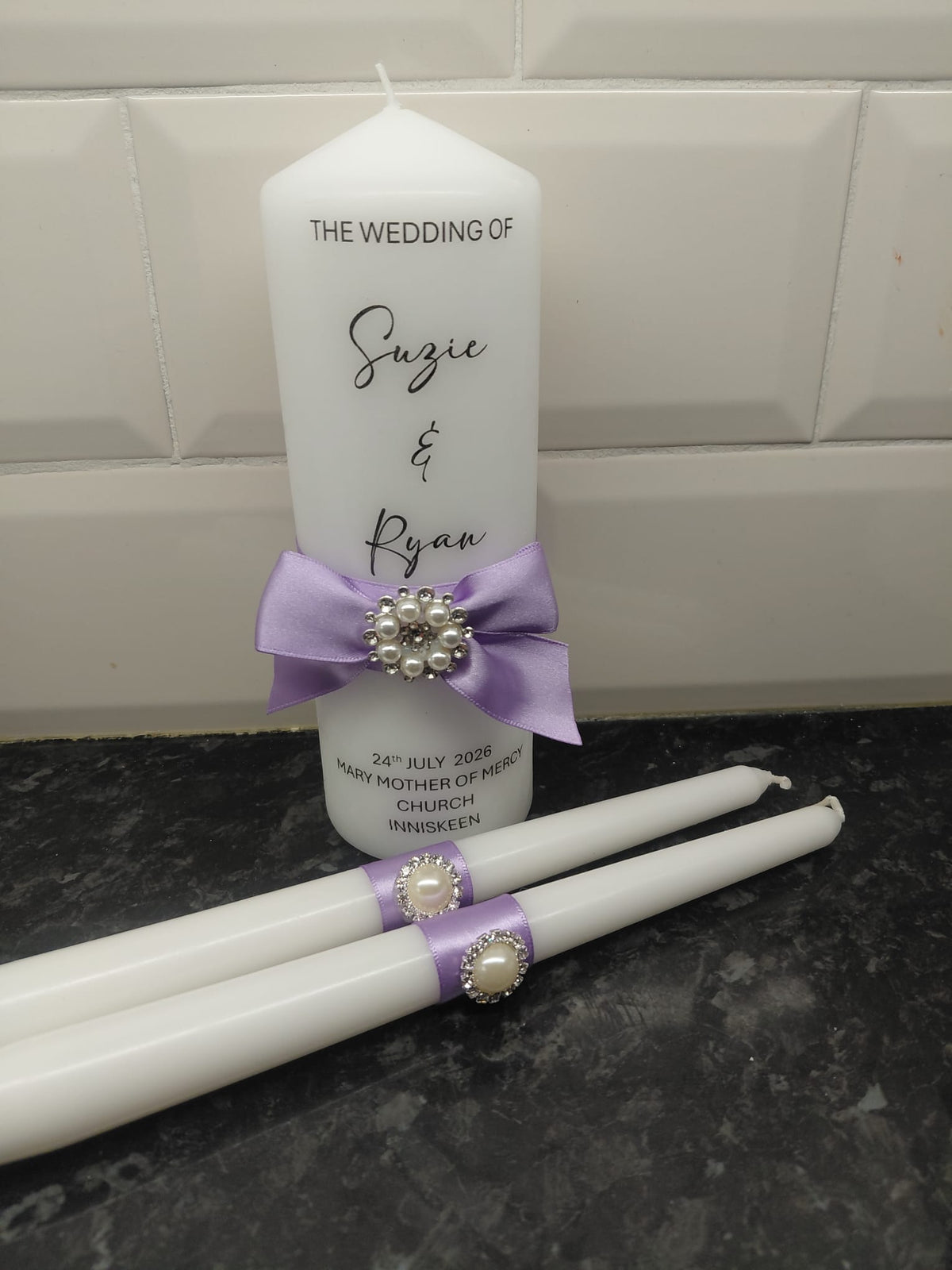 White wedding candle set with a tall pillar candle and two taper candles, decorated with light purple ribbons and pearl-like brooch accents, set against a white tiled wall and dark countertop.