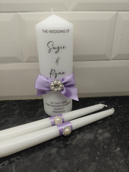 White wedding candle set with a tall pillar candle and two taper candles, decorated with light purple ribbons and pearl-like brooch accents, set against a white tiled wall and dark countertop.